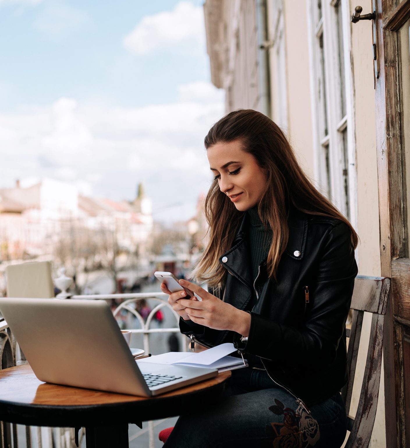 Young woman working on a balcony. Using smartphone and laptop computer outdoors.